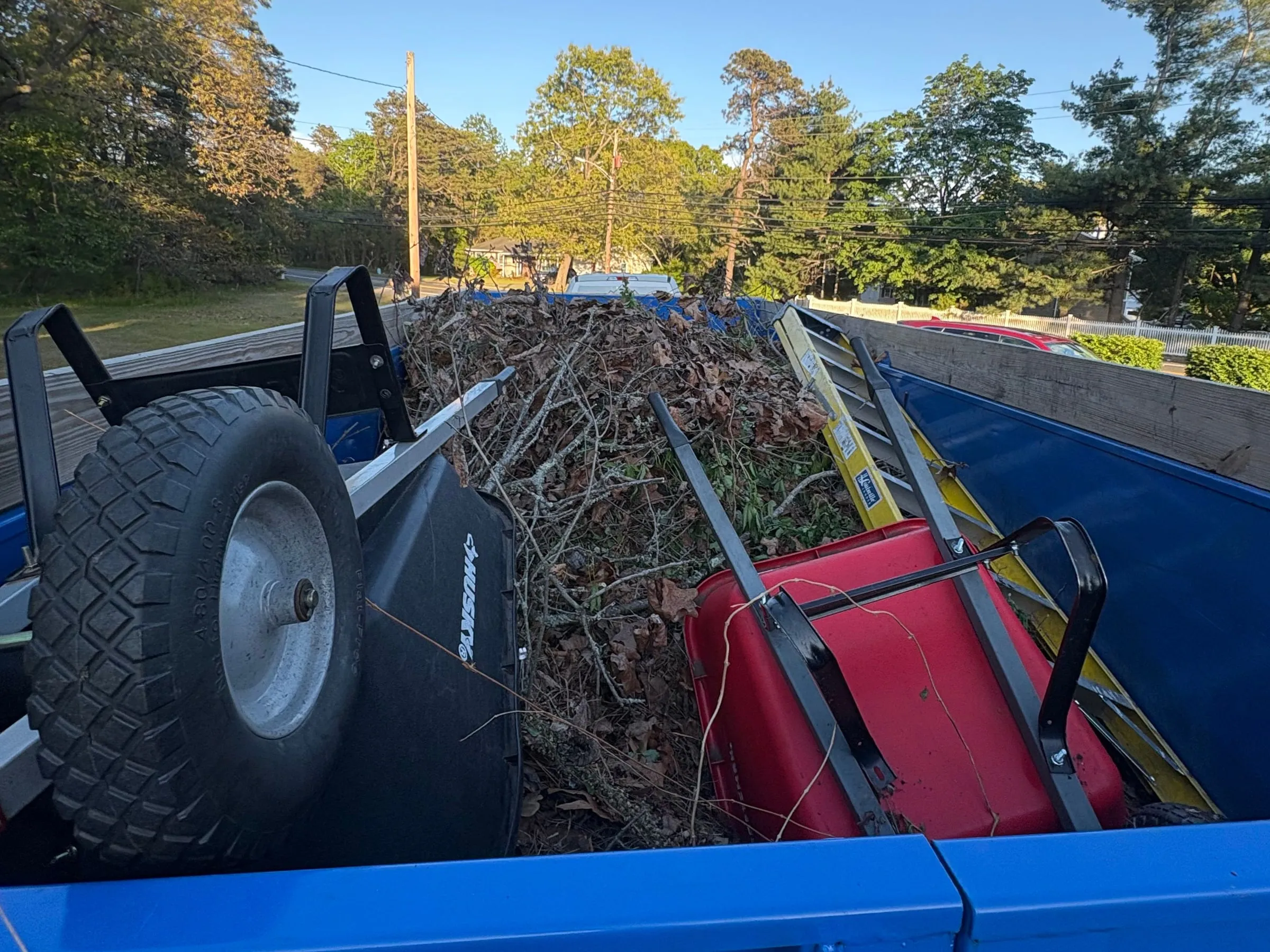 Blue dump trailer loaded with brush and leaves, two upturned wheelbarrows and a yellow ladder, with trees behind