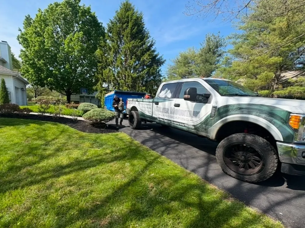Silver Mountain Landscaping wrapped pickup and blue dump trailer in a driveway beside a green lawn and mulched bed