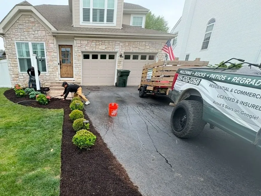 Crew finishing a new boxwood bed with dark mulch along a driveway, work truck and trailer parked alongside