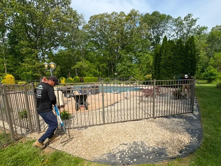 Crew member raking tan decorative stone over landscape fabric around a bronze pool fence