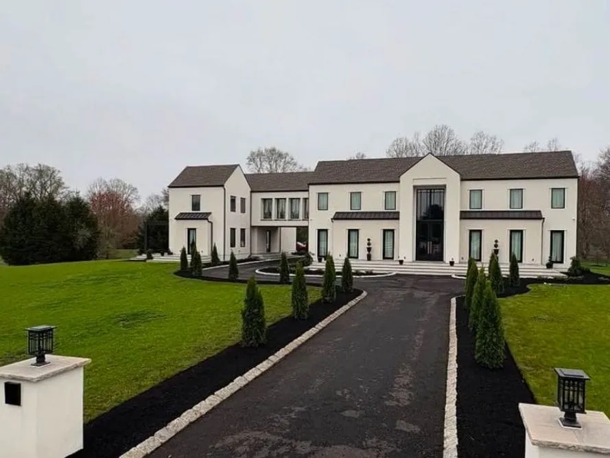 After: The same driveway lined with black mulch beds, stone edging and arborvitae leading to the white house, lantern pillars in front
