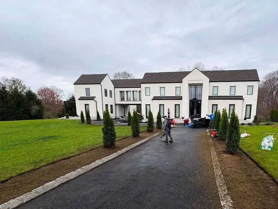 Before: Driveway with bare soil beds and newly planted arborvitae on both sides, crew working near a white house