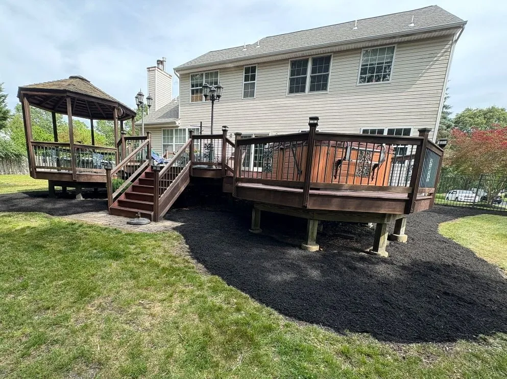 Fresh black mulch bed with a curved edge around a raised wooden deck and gazebo behind a beige two-storey house