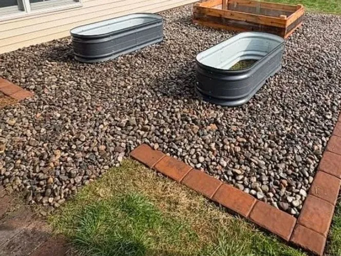 After: River rock bed edged with red pavers beside a house, holding two grey metal planter tubs and a cedar raised bed.