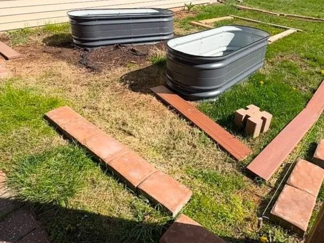 Before: Two grey metal planter tubs on patchy lawn beside a house, with red pavers and boards laid out to mark a new bed.