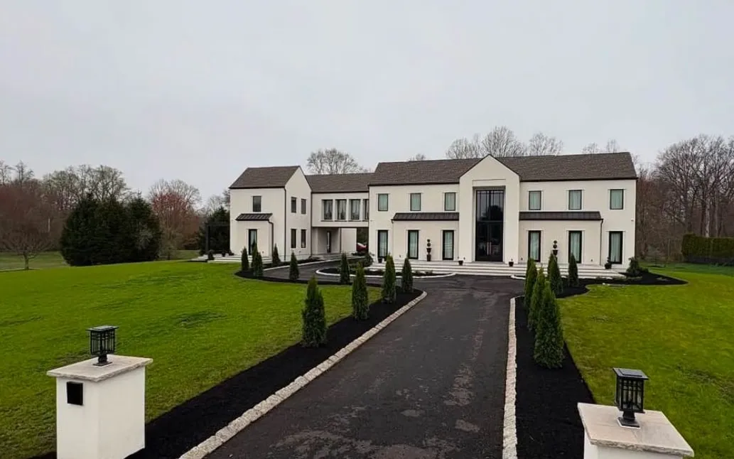 Asphalt driveway edged with cobblestone, black mulch beds and young arborvitae leading to a large white house.