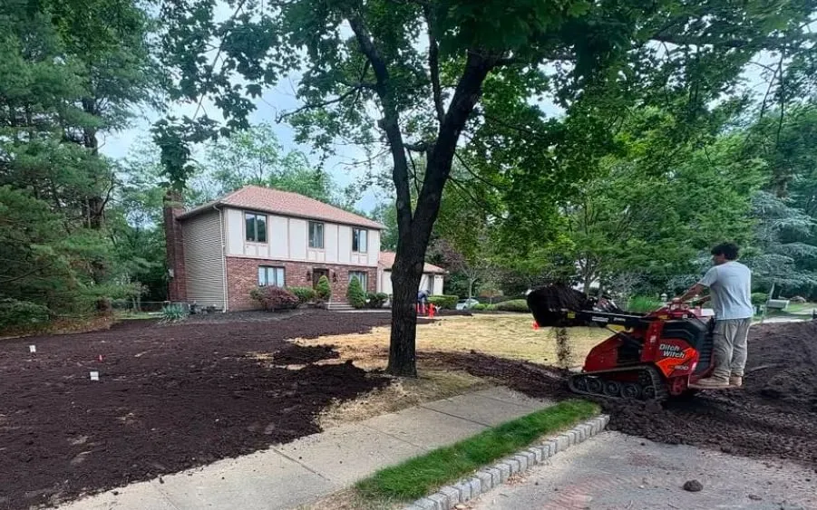 Worker on a red mini skid steer spreading dark topsoil across a front lawn, with a two-storey brick house behind