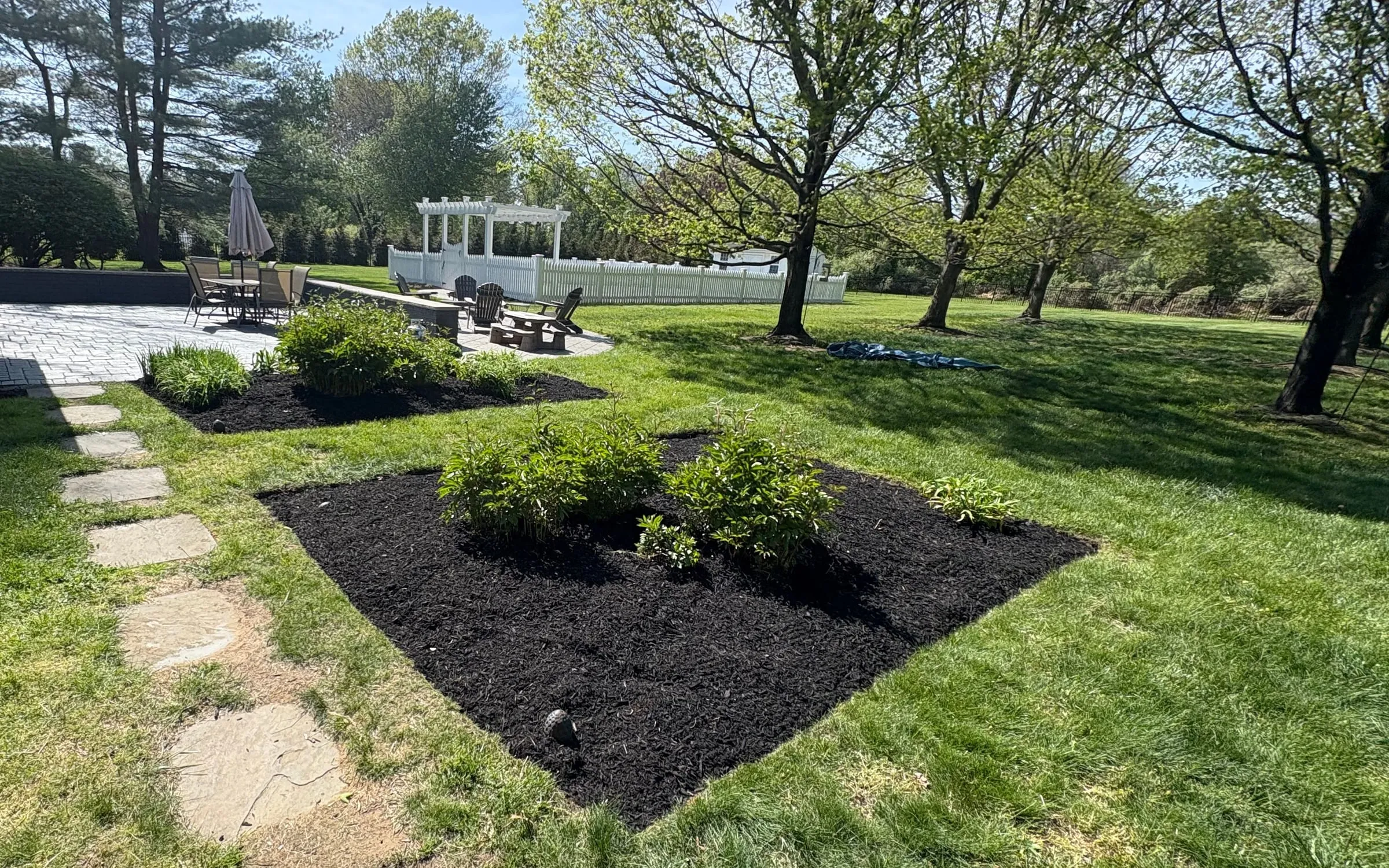 Backyard with two freshly mulched, sharply edged planting beds, a stepping-stone path, paver patio and white pergola