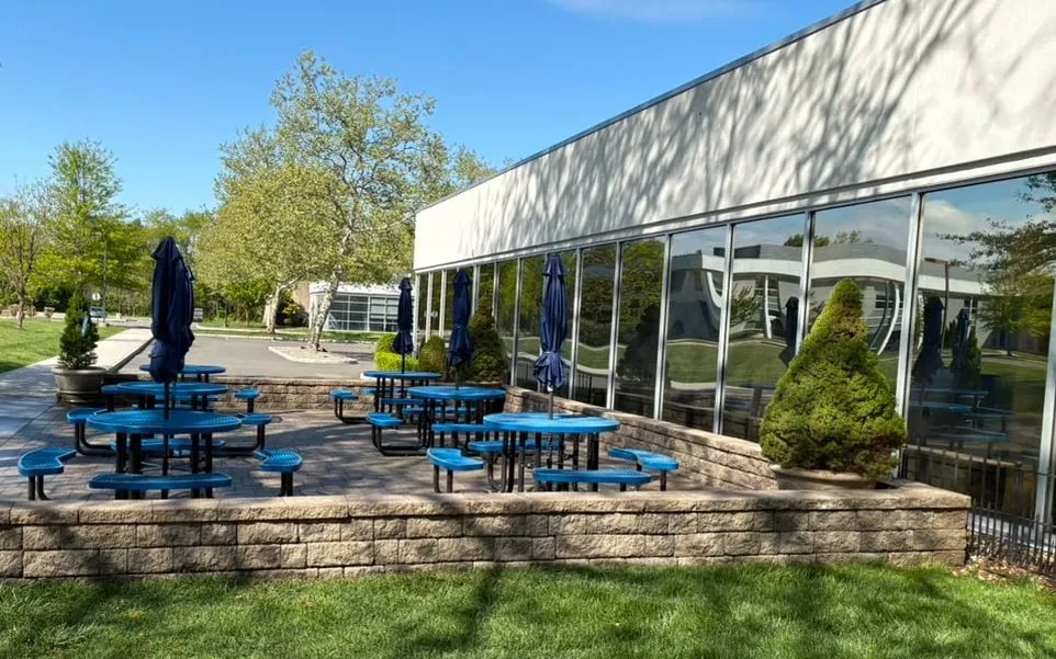 Clean tan block retaining wall around a paver patio with blue picnic tables beside a glass-fronted commercial building.