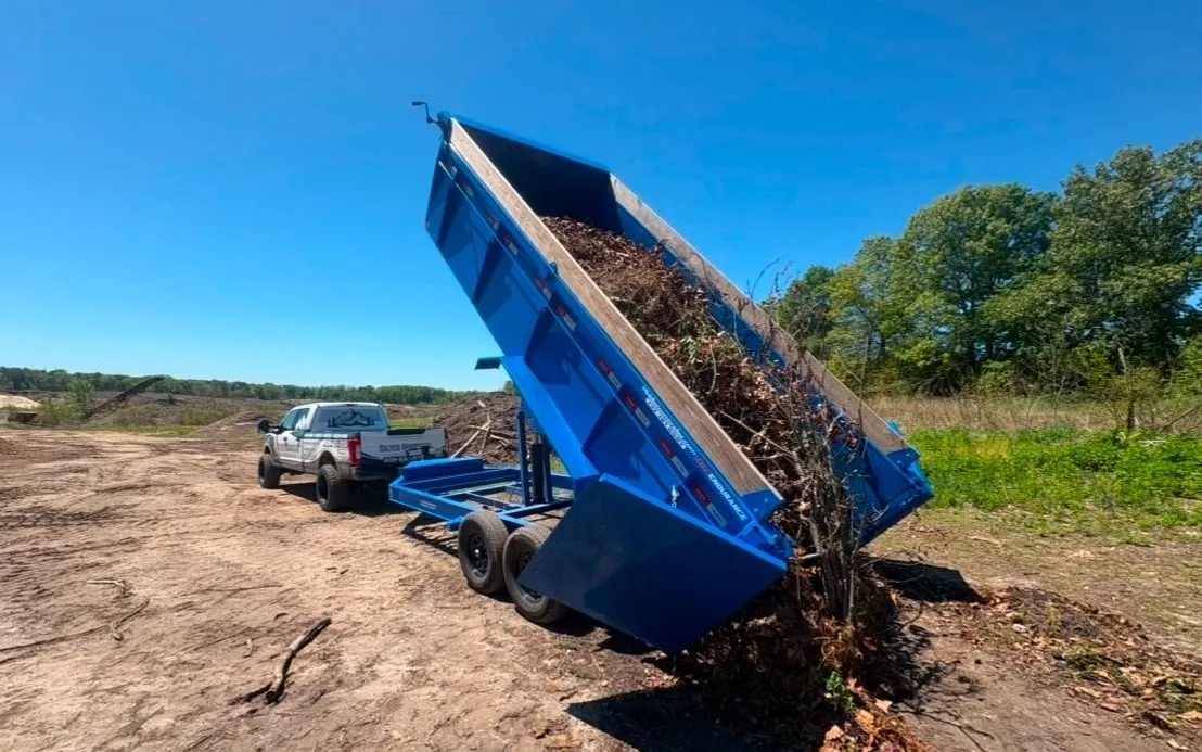 Blue dump trailer tipped up behind a white pickup, unloading brush and branches at a dirt yard-waste site.