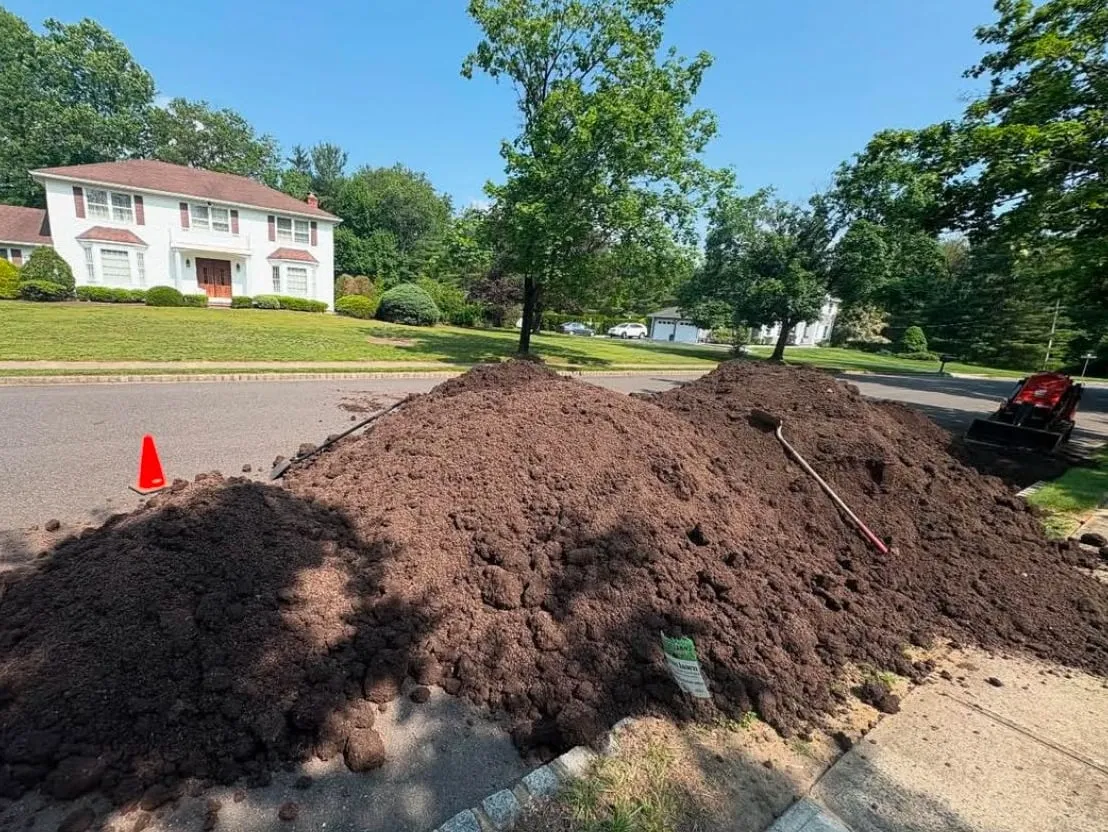 Large pile of dark topsoil delivered at the curb with a rake and shovel on it and a mini skid steer nearby